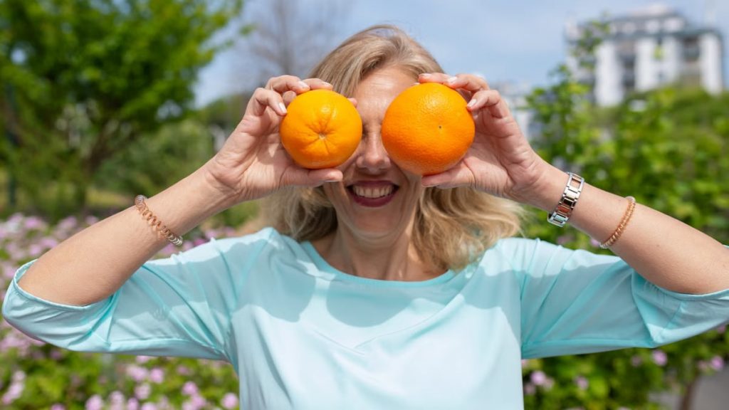 A smiling woman playfully holding oranges in a sunny park setting.